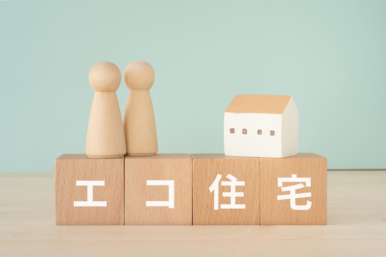 木のブロックが4つ横に並んでいる。ボックスには1文字づつエコ住宅と書かれている。ボックスの上に人形が2体と家が乗っている。