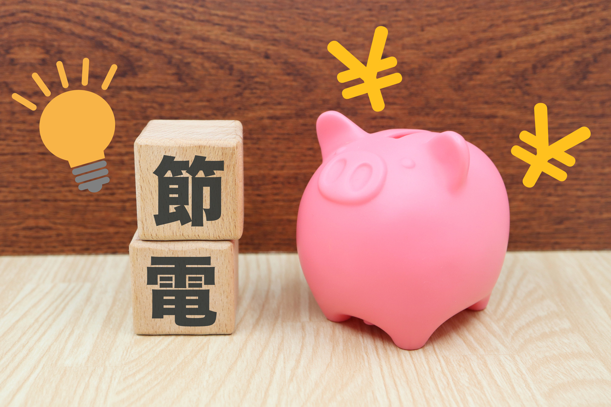 木のボックスが2つ積まれている。木のボックスには節電と書かれている。右側には豚の貯金箱と円マークがある画像。