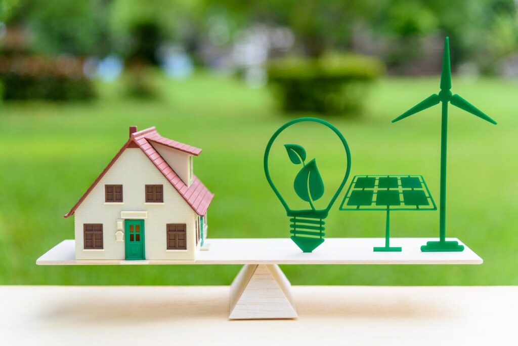 緑の自然な背景でシーソーの上に一軒家と電球、ソーラーパネル、風力発電のイラストが乗っている絵が描かれたイラスト