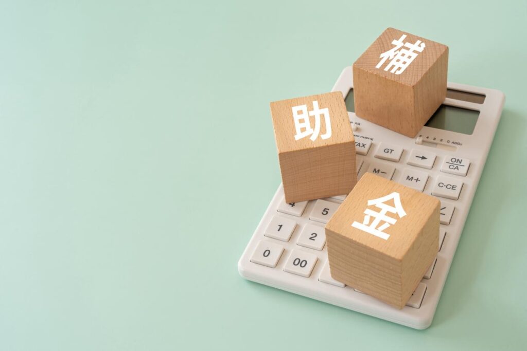 薄い緑の背景に電卓、電卓の上に補助金と書かれた木のブロックが乗っている画像