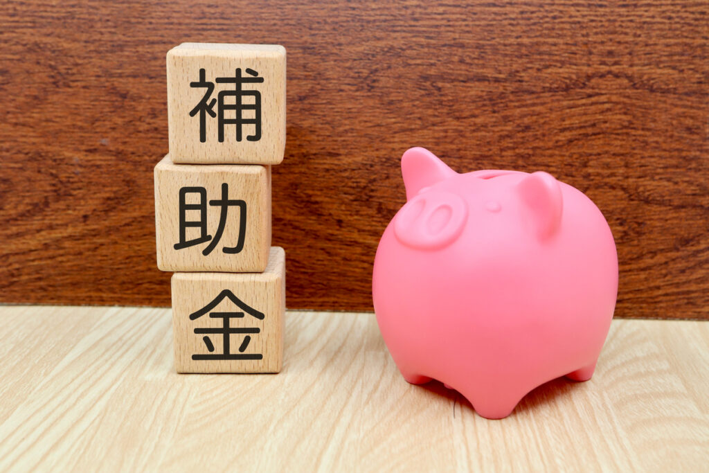 木のブロックが左側に3つ積み上げられている。上から読むと補助金。右側には豚の貯金箱が置かれている画像