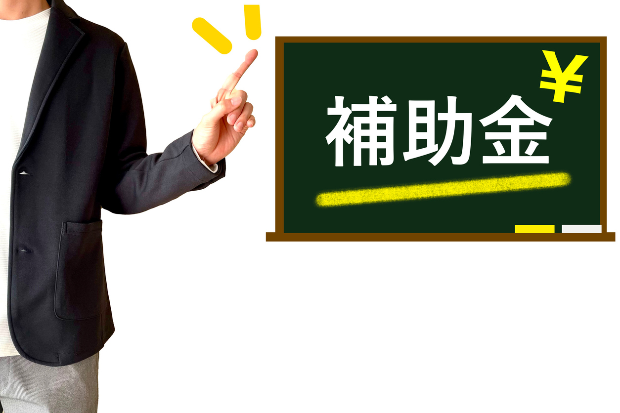 ジャケットを着た男性が黒板を指差している画像。黒板には補助金と書かれている