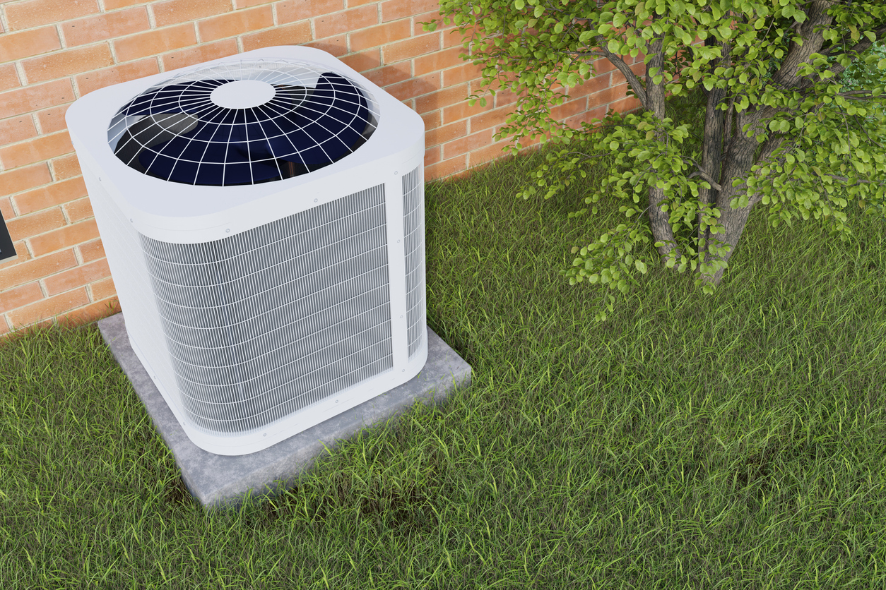 屋外の芝生の上に設置された室外機。レンガの壁の近くにあり、隣には木が植えられている画像