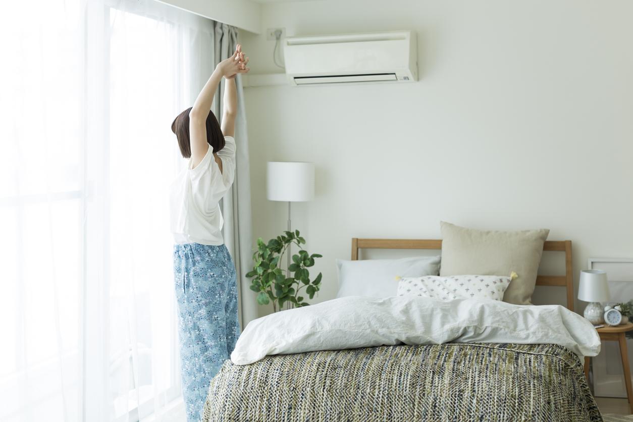 明るい寝室で、女性がカーテンの前で伸びをしている様子。エアコンや観葉植物、ベッドが映る朝の風景画像