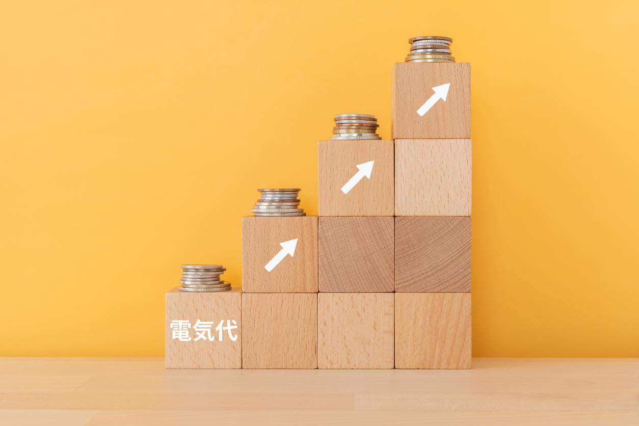 黄色の背景に、階段状に積み重ねられた9つの木製ブロックがある。一番下の左端のブロックには日本語で「電気代」と書かれており、その上の各ブロックには白い上向きの矢印と、その上に積み重ねられた硬貨が配置されている画像