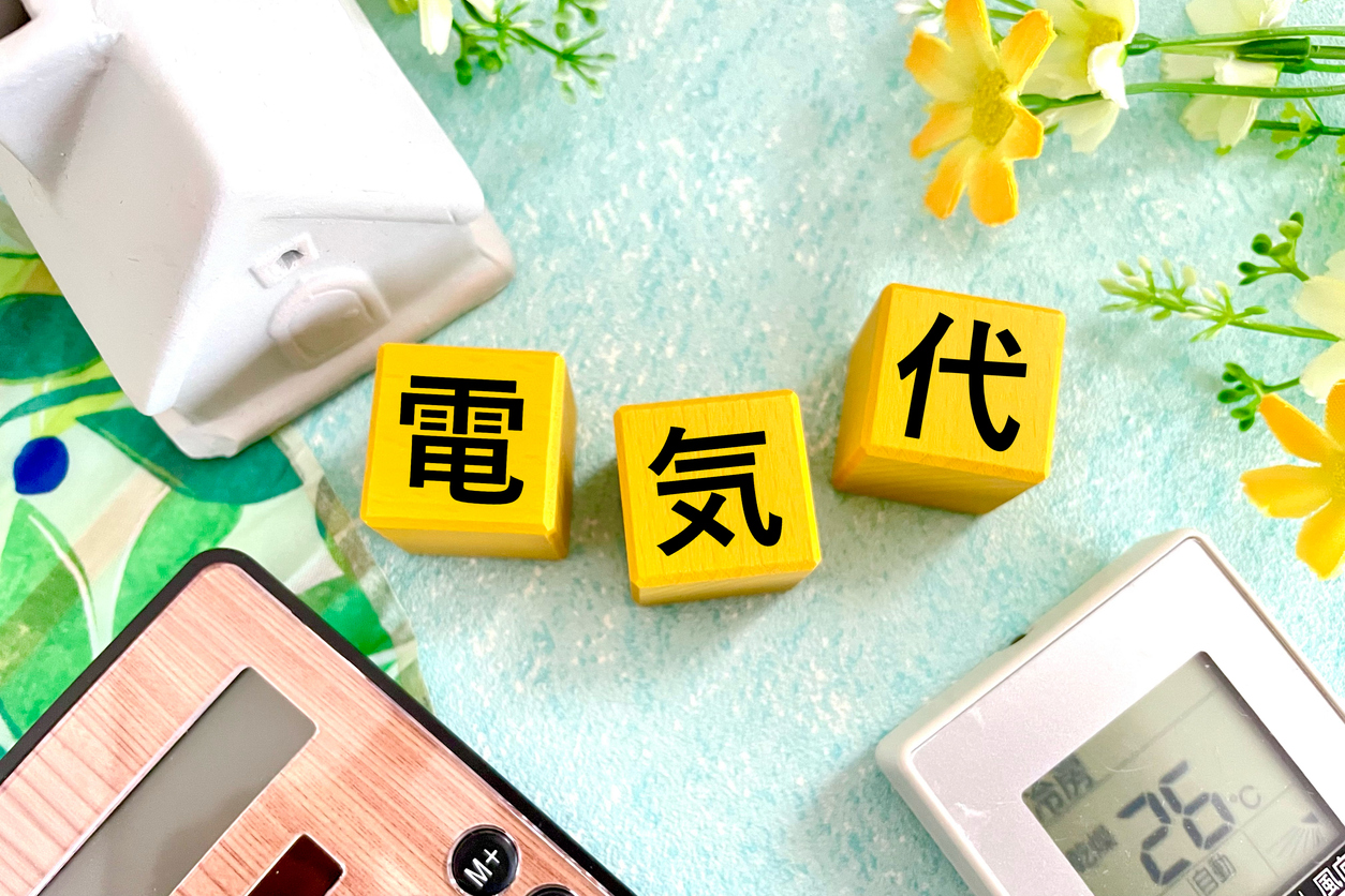 「電気代」と書かれた黄色いブロックが並び、周囲に家の模型、電卓、温度計(26℃表示)、花が配置されている画像