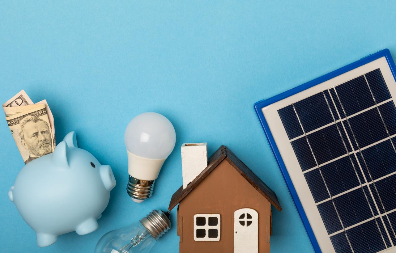 青背景の上に、青い豚の貯金箱、紙幣、電球、家の模型、太陽光パネルが並んでいる画像