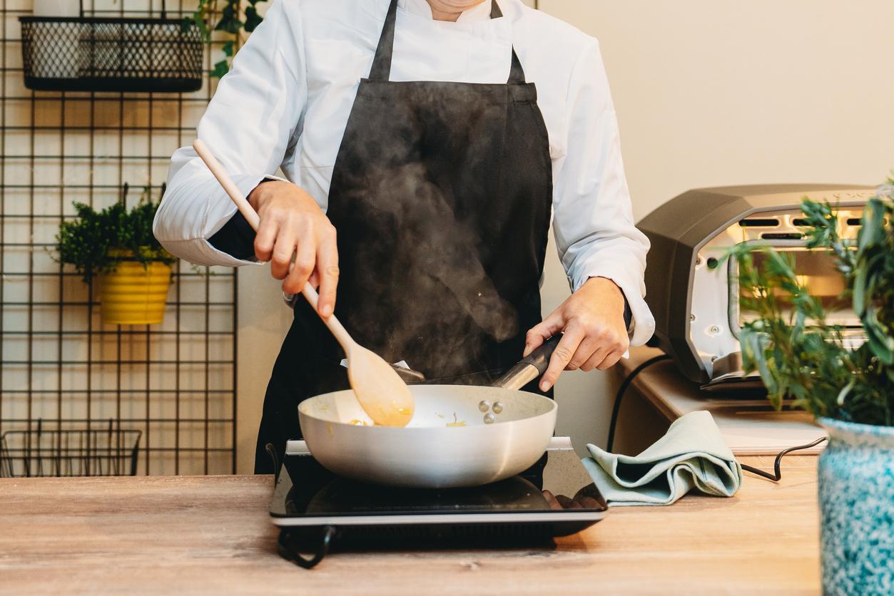 黒いエプロンを着た料理人が、卓上IHクッキングヒーターでフライパンを使い調理している様子。湯気が立ち上り、手元には木べらとナプキンが置かれている画像