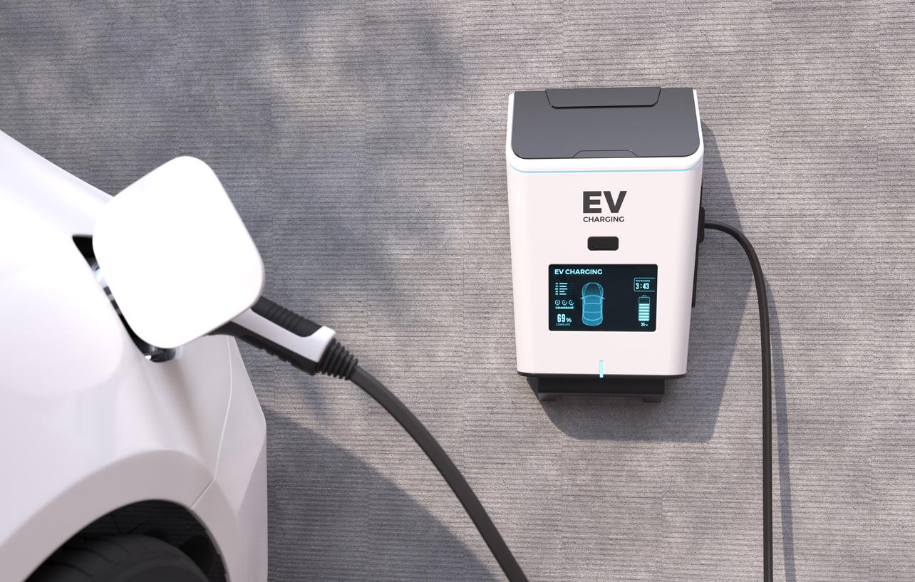 白い電気自動車が壁に取り付けられたEV充電ステーションに接続されており、充電ステーションの画面にはEV充電のステータスが表示されている画像