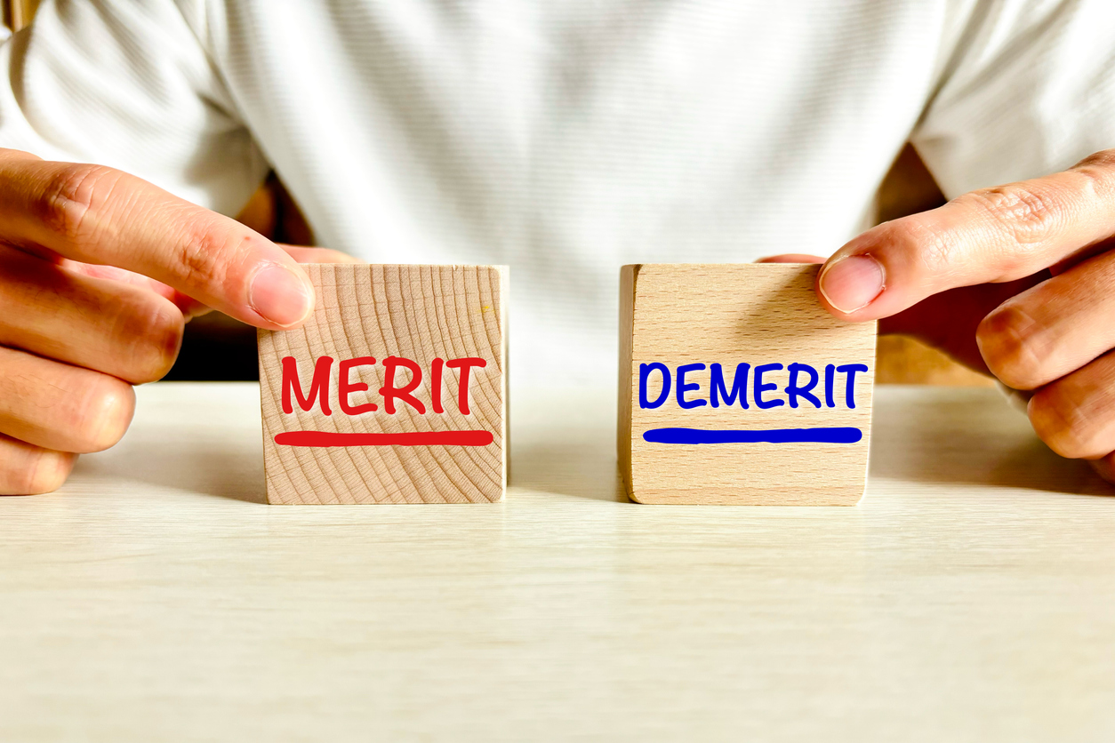 机の上に置かれた「MERIT(メリット)」と「DEMERIT(デメリット)」と書かれた木のブロックを、それぞれ左右の手で掴んでいる画像